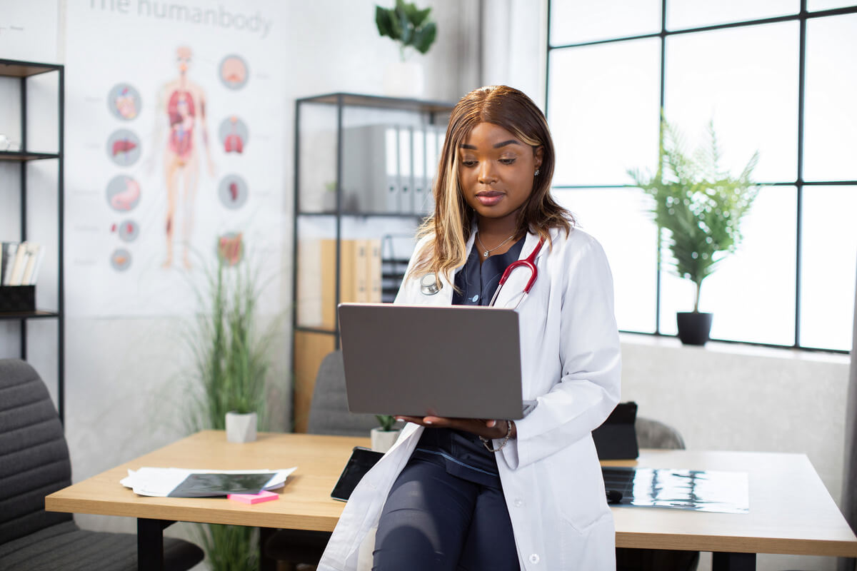 A healthcare professional leans against a desk and looks at a laptop, representing the idea of creating educational content for healthcare websites.