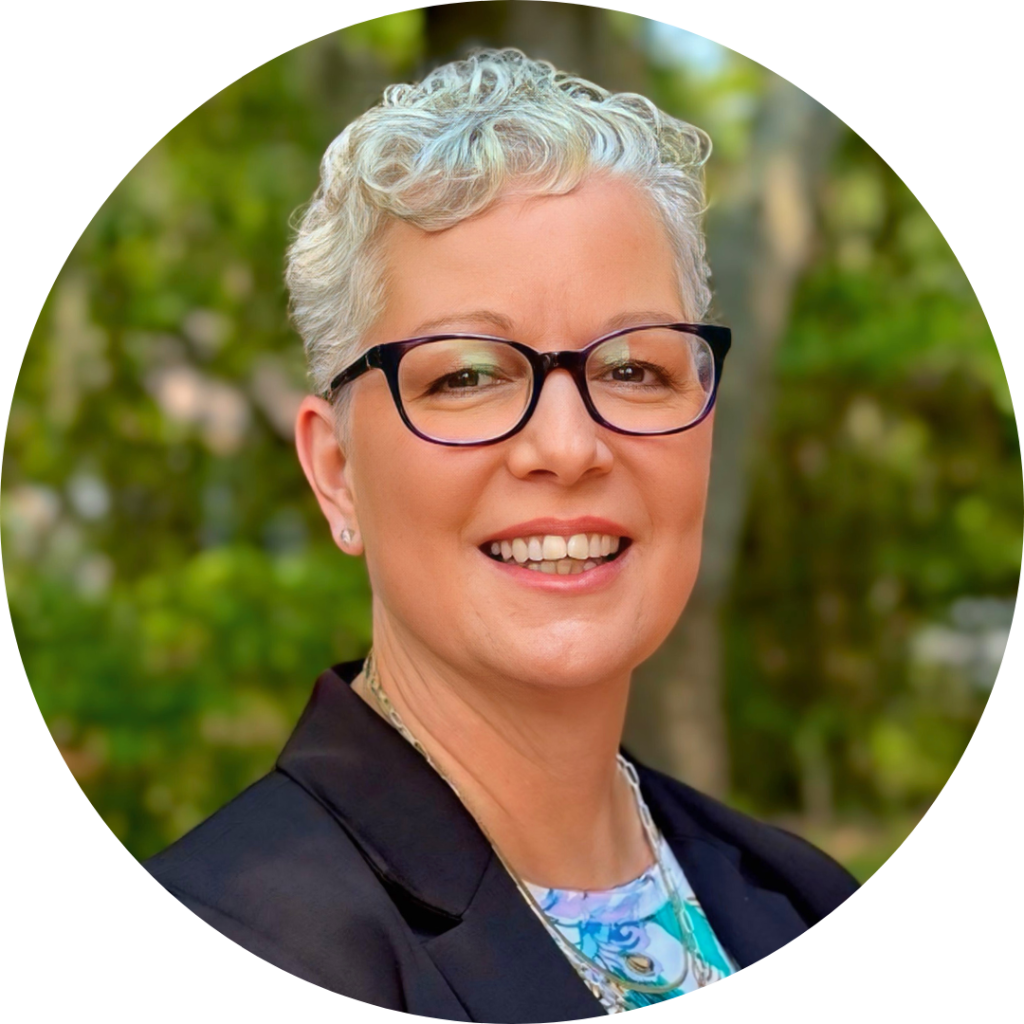 Marcell Mallette, Winkler Group Consultant, smiles at the camera. She has curly silver hair and rimmed black glasses. She's wearing a navy-blue suit jacket with a patterned blue blouse underneath,
