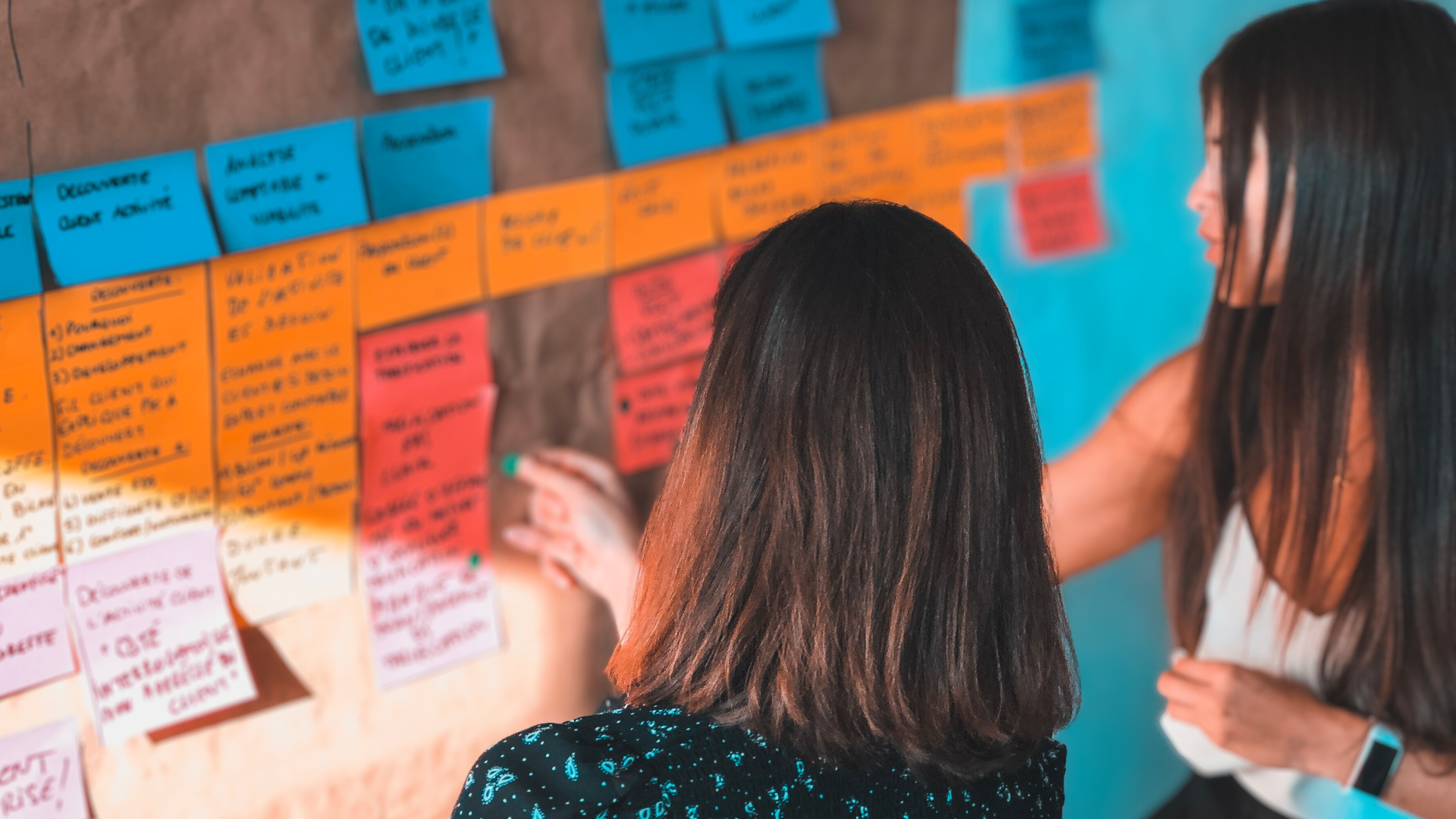Header image for "Three Conversations Every Nonprofit Board Should Have Before Year-End Giving Season." Two women, with their backs turned to the camera, work on a corkboard full of brightly colored sticky notes.
