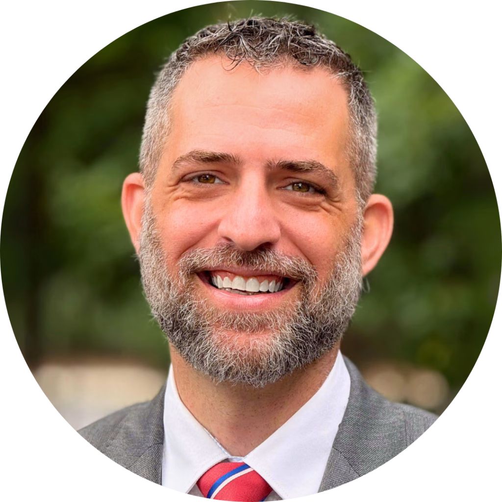 Josh Olmsted, Winkler Group Consultant, smiles at the camera. He has short hair and a cropped salt-and-pepper beard with a grey suit jacket, white dress shirt, and red and blue striped tie.