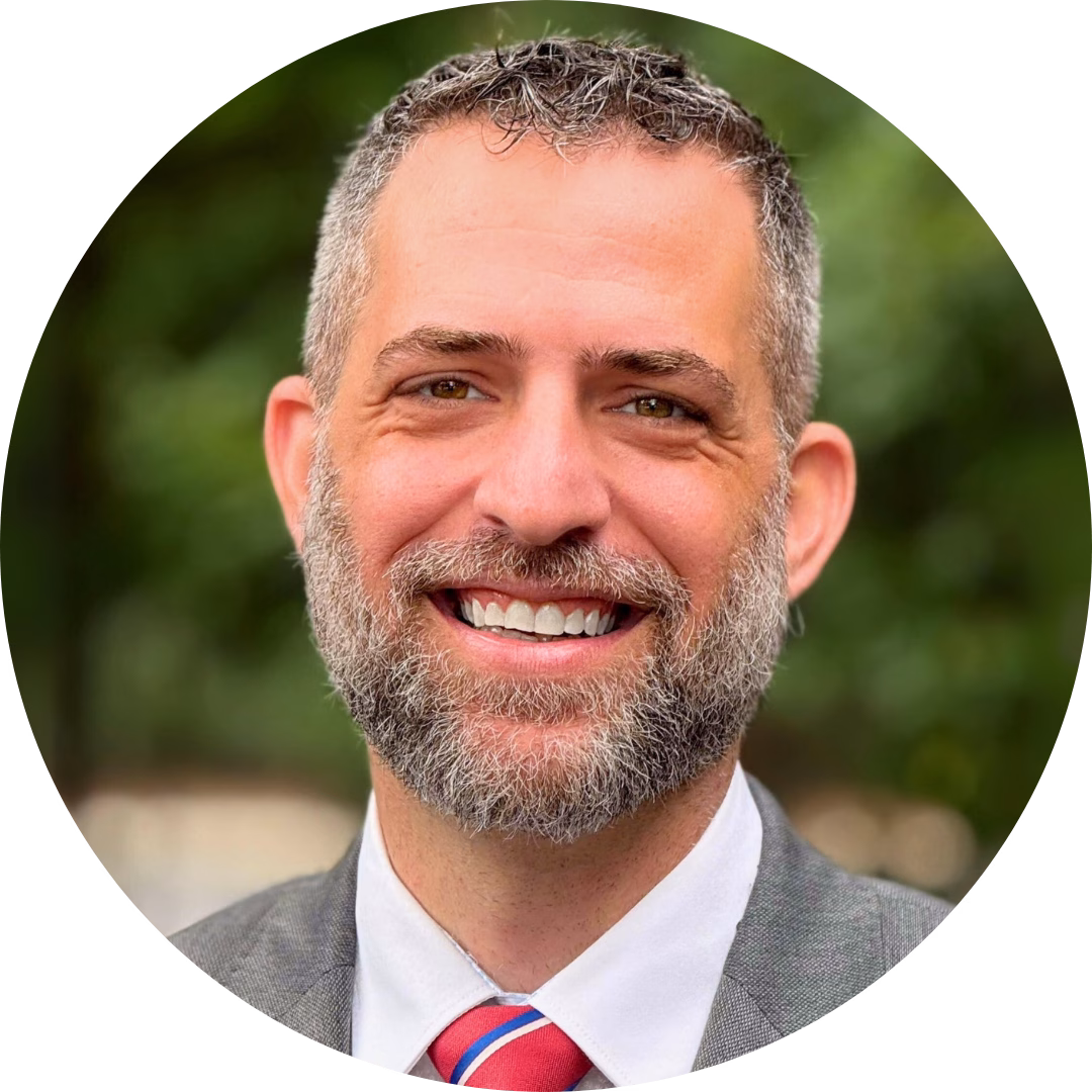 Josh Olmsted, Winkler Group Consultant, smiles at the camera. He has short hair and a cropped salt-and-pepper beard with a grey suit jacket, white dress shirt, and red and blue striped tie.